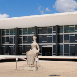 Sede do Supremo Tribunal Federal, edifício suspenso próximo ao chão com colunas modernas brancas e caixa de vidro escuro, com estátua da justiça sentada à frente.