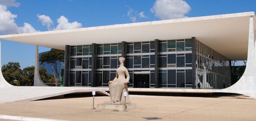 Sede do Supremo Tribunal Federal, edifício suspenso próximo ao chão com colunas modernas brancas e caixa de vidro escuro, com estátua da justiça sentada à frente.
